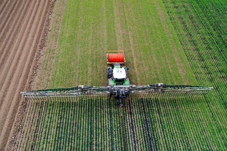 Aerial view of a tractor spreading slurry on crops with wide booms across a green field divided into sections.