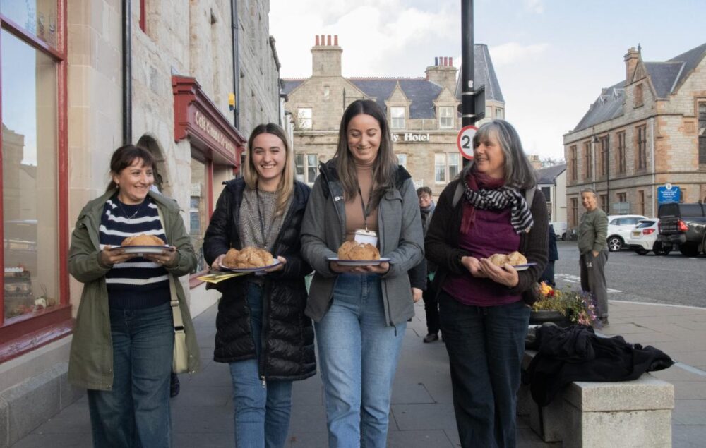 four women walking in an urban setting carrying a plate of food each