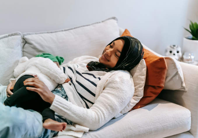 Woman in black hijab lying on a white couch with a baby on her tummy