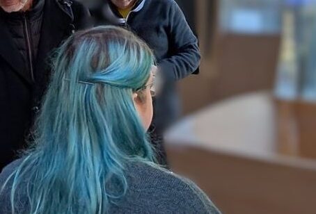 Person with long blue hair and a dark sweater stands inside a wooden workshop or studio with blurred figures in the background.