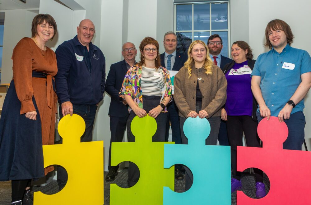 Group of eight people standing inside holding large colourful puzzle pieces in yellow, green, blue, and red.