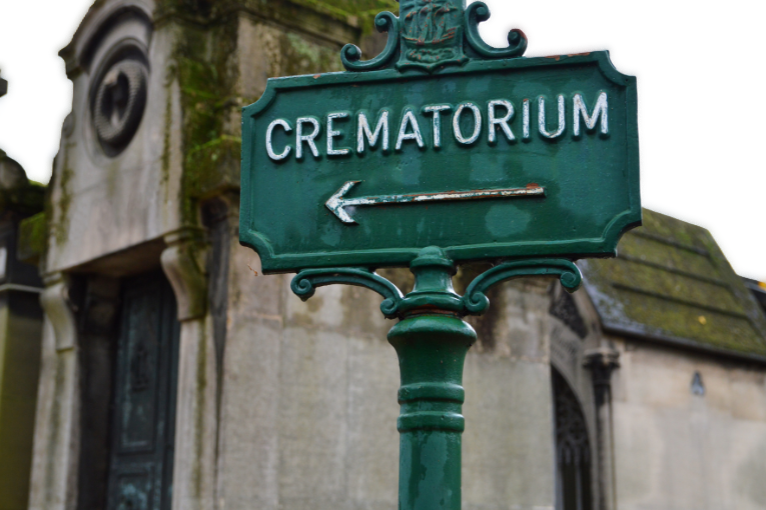 Green metal signpost with the word 'CREMATORIUM' and a left-pointing arrow in front of old stone mausoleums.