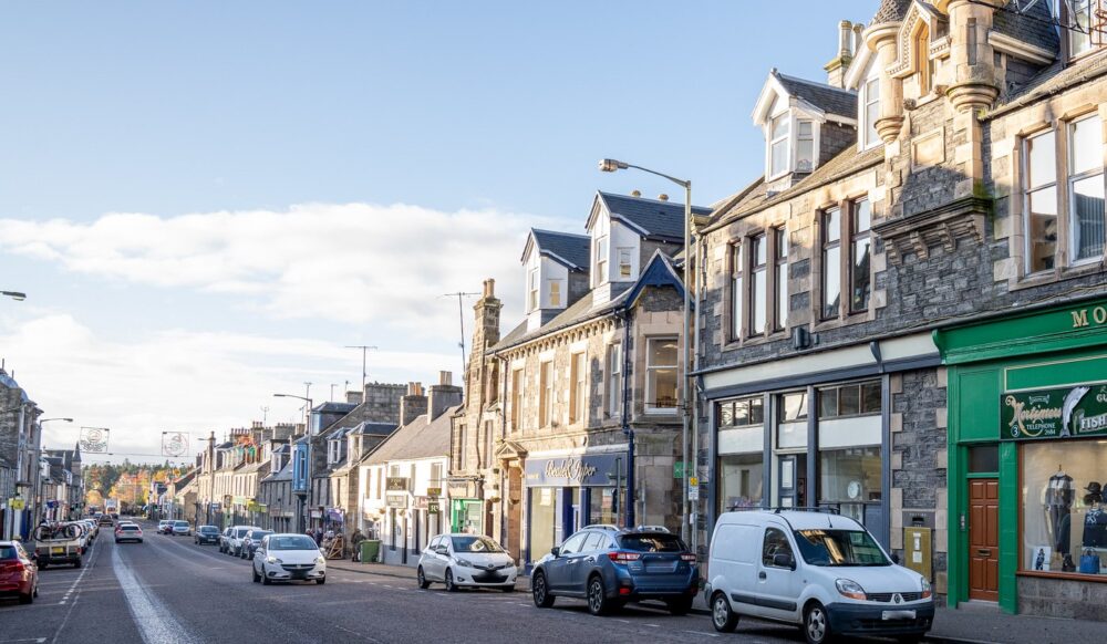 Historic stone buildings line a busy street with parked cars under a clear blue sky in a small town.