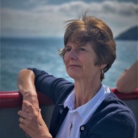 Woman with short hair wearing a navy cardigan and white shirt, sitting on a boat with ocean and distant hills in the background.
