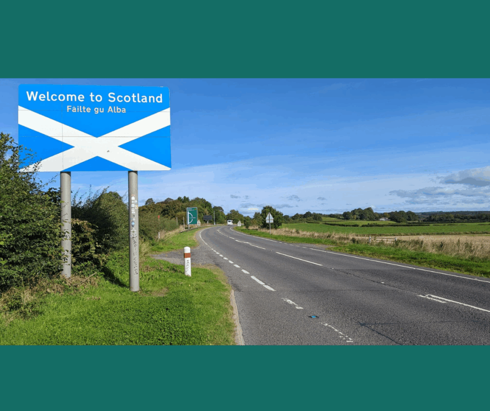 Scenic road leading into Scotland marked by a large blue welcome sign with the Saltire flag.