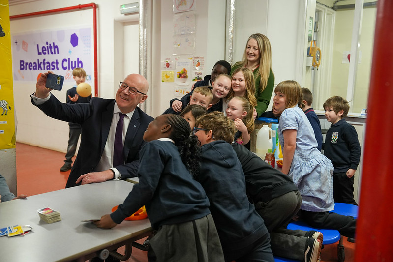 First Minister, John Swinney, takes a selfie with a group of smiling children gathered around a table at Leith Breakfast Club.