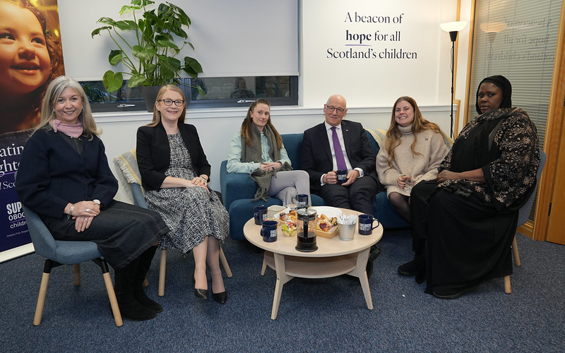 Group of six adults, including First Minister and Cabinet Secretary for Social Justice, sitting on chairs and a sofa in a meeting room with a plant and motivational poster in the background.