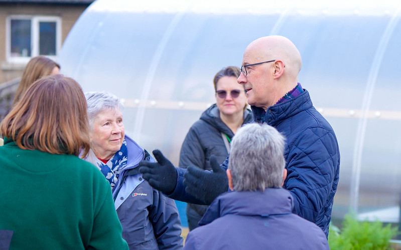 Group of adults engaged in an outdoor conversation near a greenhouse on a chilly day.