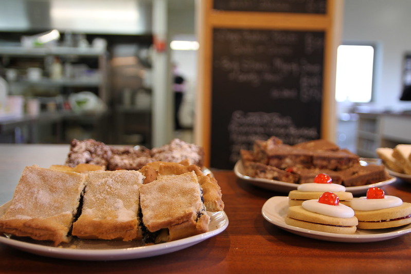 Close-up of assorted baked goods including cherry-topped biscuits and fruit cakes on plates inside a farm shop café.