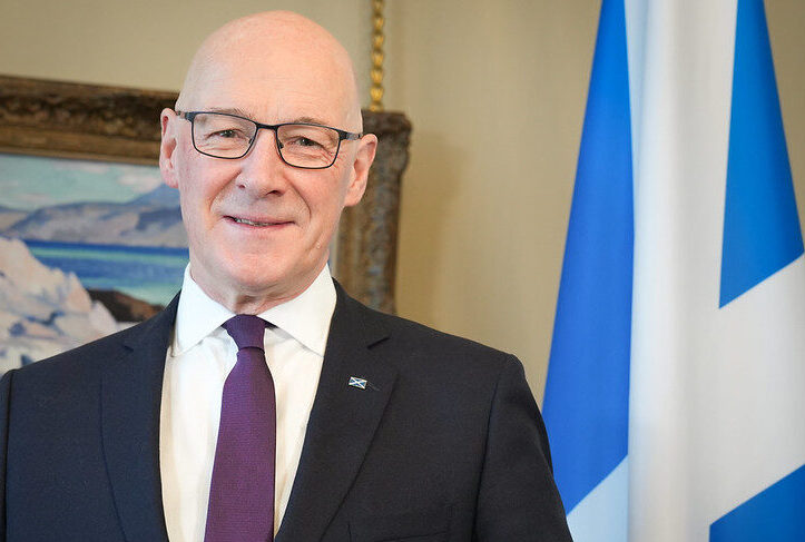 First Minister of Scotland, John Swinney, dressed in a dark suit and a purple tie standing in front of a Saltire flag and a framed painting.