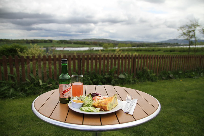Outdoor table with a plate of savoury pie, salad, a bottle of cider, and a glass of juice against a countryside backdrop.