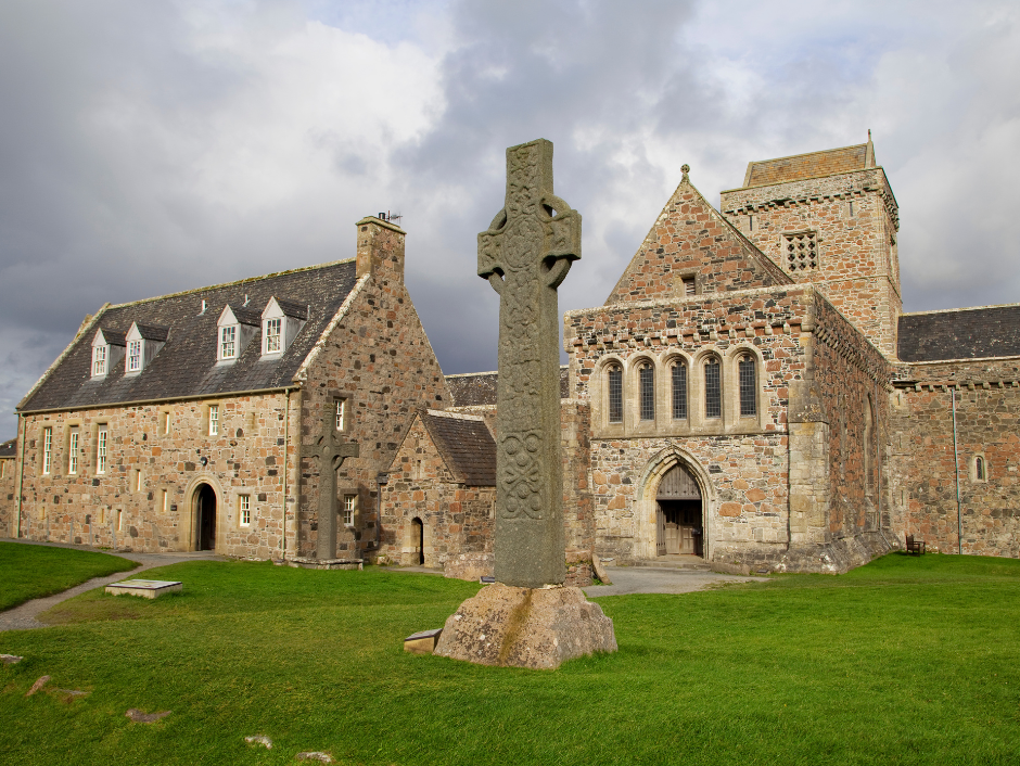 Ancient stone Celtic cross standing on green grass in front of historic medieval stone abbey in Iona, Scotland, under a cloudy sky.