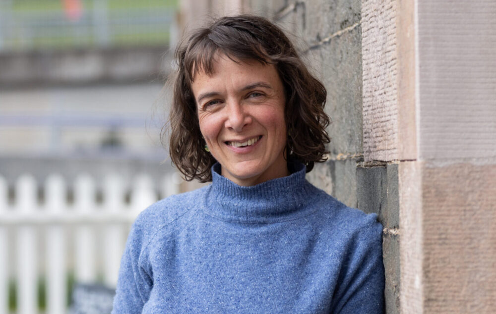 Person in a blue turtleneck sweater leaning against a stone wall outdoors near a white picket fence.