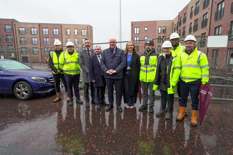 Group of construction workers and professionals standing on a wet site with residential buildings in the background.