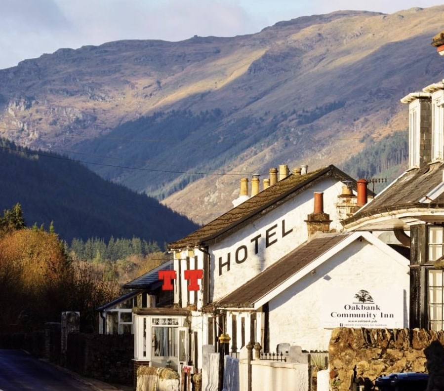White hotel building with red signage nestled at the base of sunlit, rugged mountains under a partly cloudy sky.
