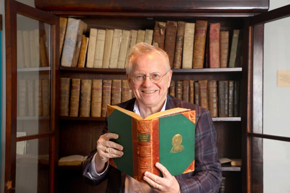 Person in a checked jacket reading a large green and brown book in front of a wooden bookshelf filled with old books.