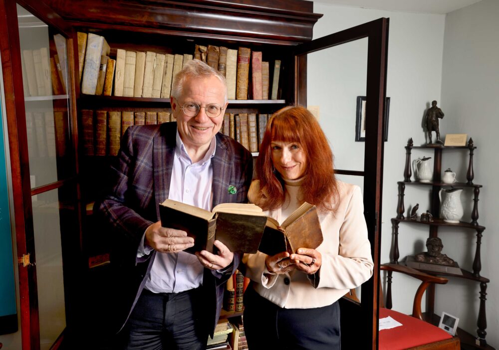 Two people standing in front of a wooden bookshelf filled with old books, each holding and reading a book.