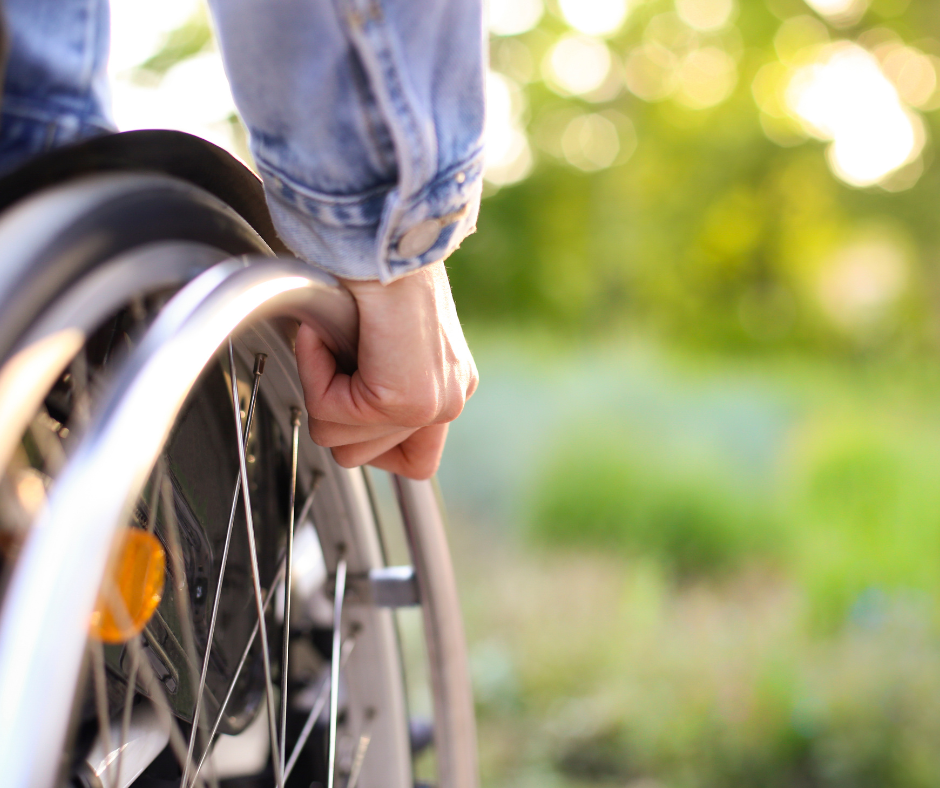 Close up image of a person in a light blue shirt with their hand on the rim of a wheelchair wheel with foliage in the bacjground
