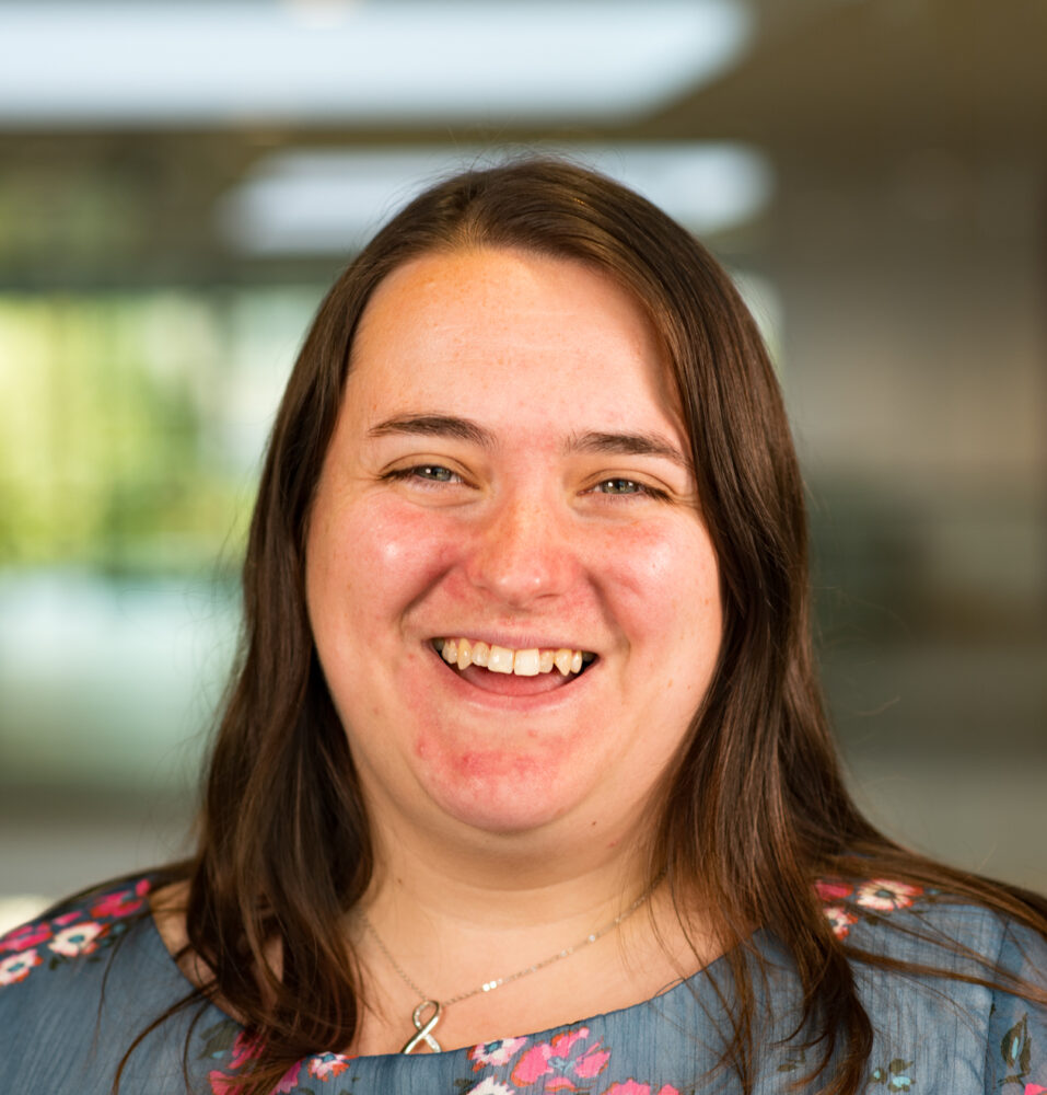 Woman with long brown hair wearing a blue floral blouse standing indoors with blurred background.