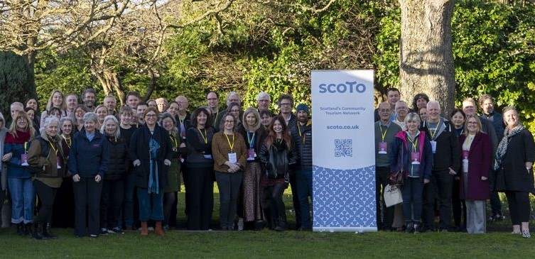 Large group of people standing outdoors in front of trees beside a SCOTO community banner on a sunny day.