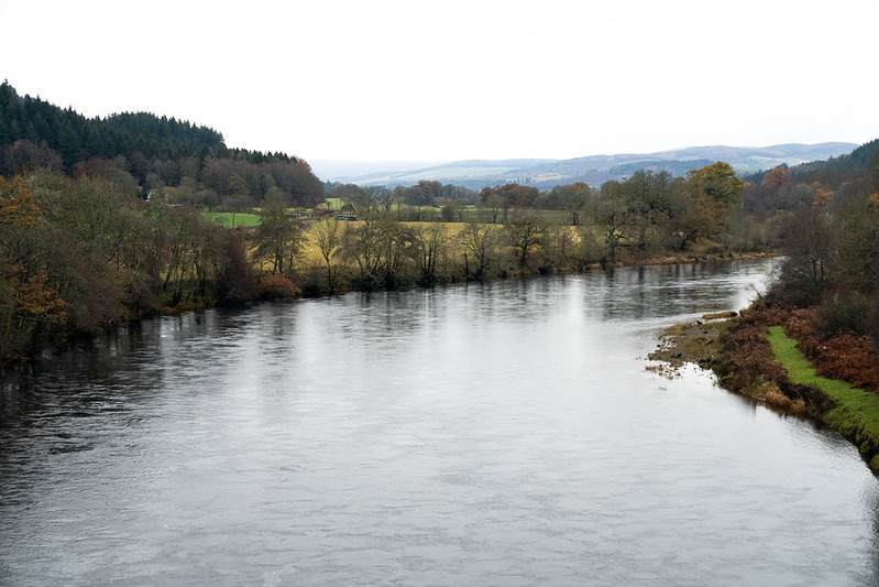 Wide river flowing gently through a rural landscape with bare trees lining its banks under an overcast sky.