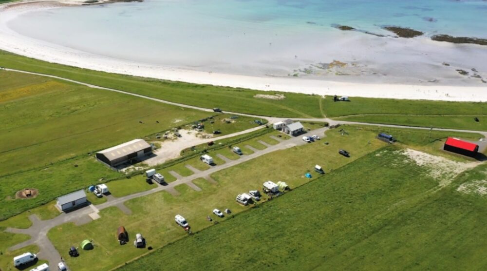 Aerial view of a coastal campsite with multiple parked campervans and tents near a white sandy beach and turquoise water.