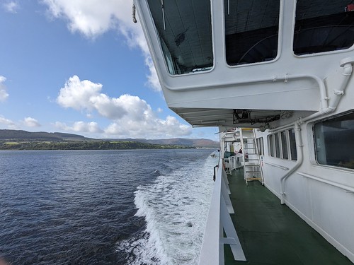 View from the side deck of a ferry showing the wake trailing behind as it moves through calm waters under a partly cloudy sky.