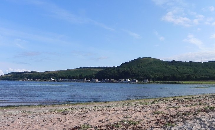 Sandy beach with sparse vegetation in the foreground and calm water leading to a green hill with scattered houses along the shoreline under a clear blue sky.