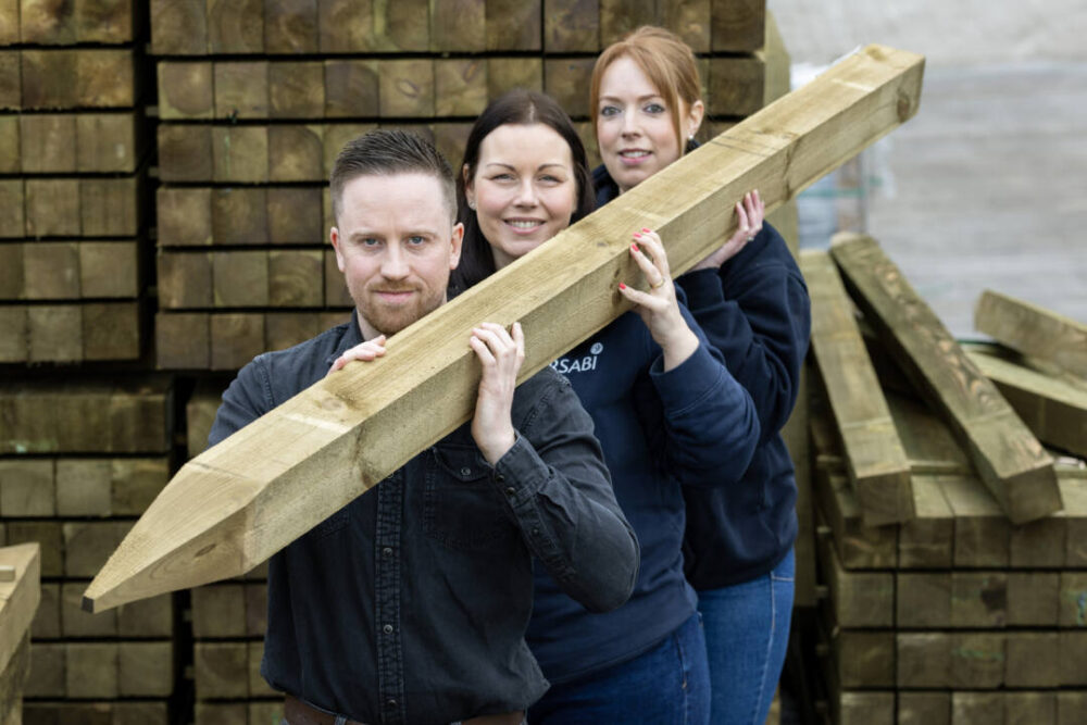 Three people standing in a row holding a large pointed wooden beam in a lumber yard with stacks of timber behind them.