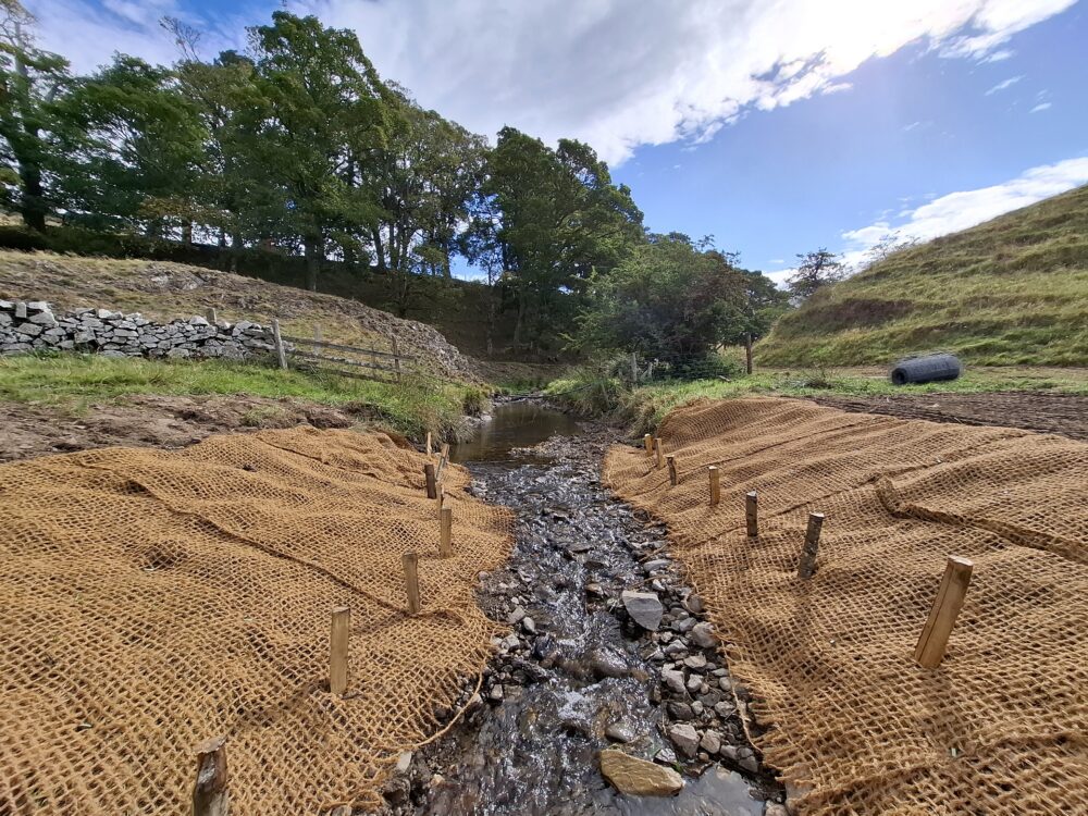 A small rocky stream flows between banks covered with erosion control mats secured by wooden stakes under a partly cloudy sky.