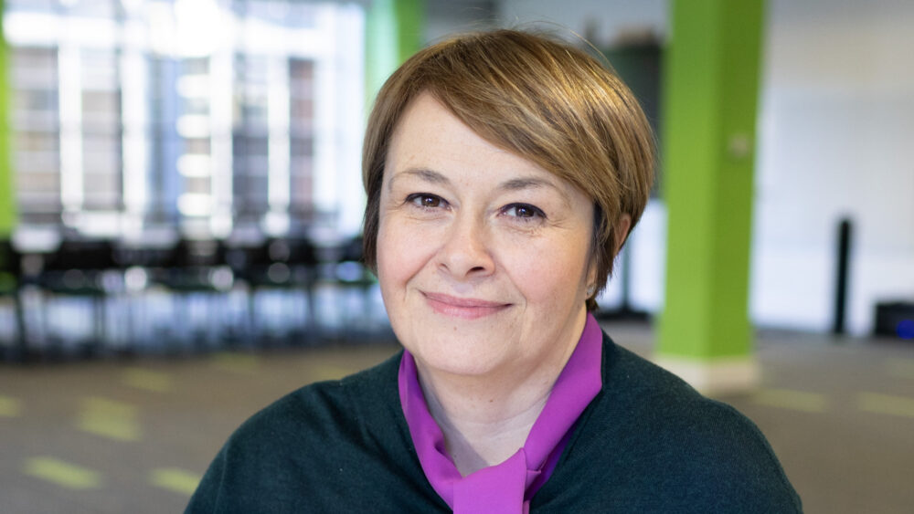 Woman with short light brown hair wearing a dark green sweater and purple collared shirt in a bright modern office space.