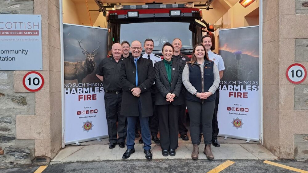 Group of firefighters and officials posing in front of a fire engine inside a fire station with safety banners on each side.