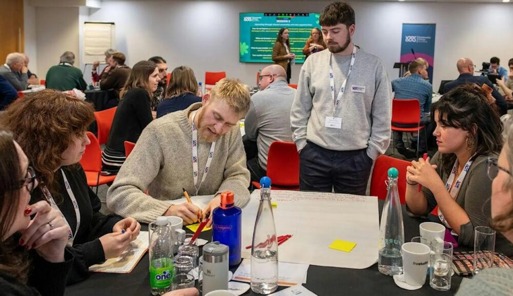 Group of professionals engaged in a collaborative discussion around a table with notes and water bottles during a conference.