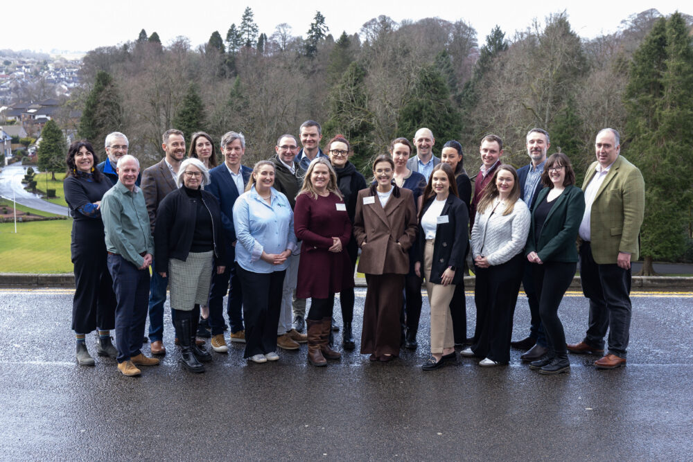 Group of 20 adults standing outdoors on a paved area with trees and a town visible in the background on a cloudy day.