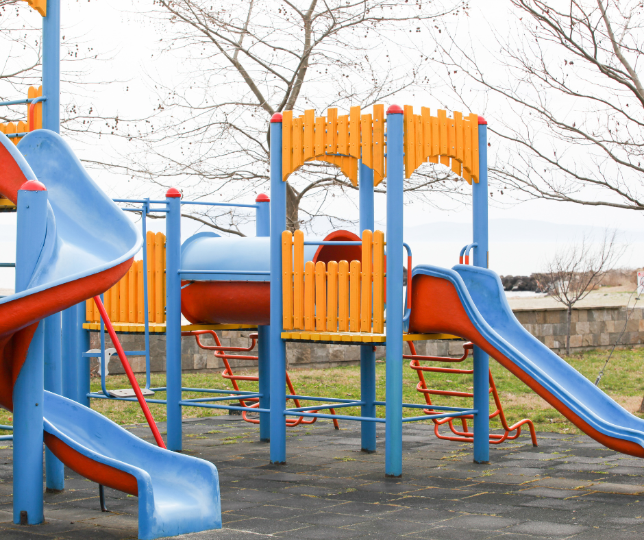Colourful playground structure with blue slides, orange railings, and climbing ladders on a rubber mat surface under leafless trees.