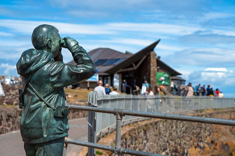 Bronze statue of a person with binoculars overlooking a busy observation deck under a blue sky.