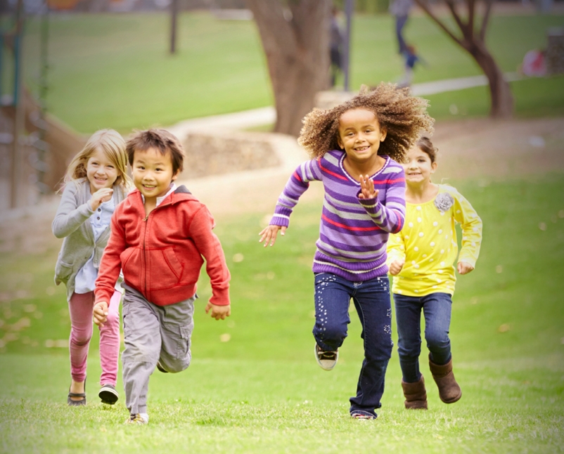 Four children running energetically across a grassy park on a bright day, with trees and pathways in the background.