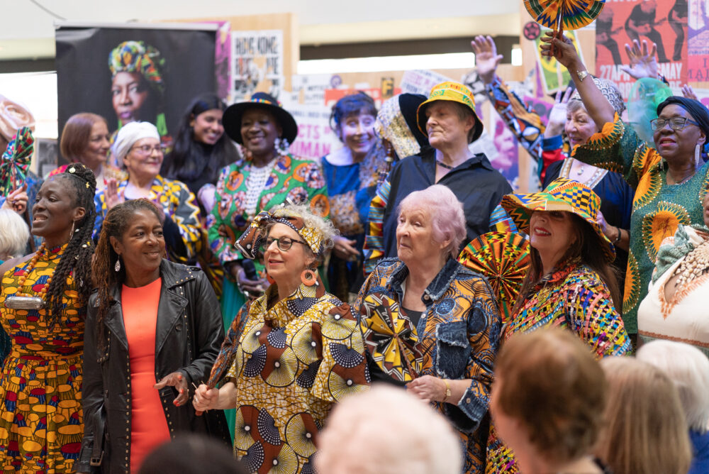 Group of people wearing colourful African print clothing gathered indoors, some holding matching fans, engaged in a lively event.