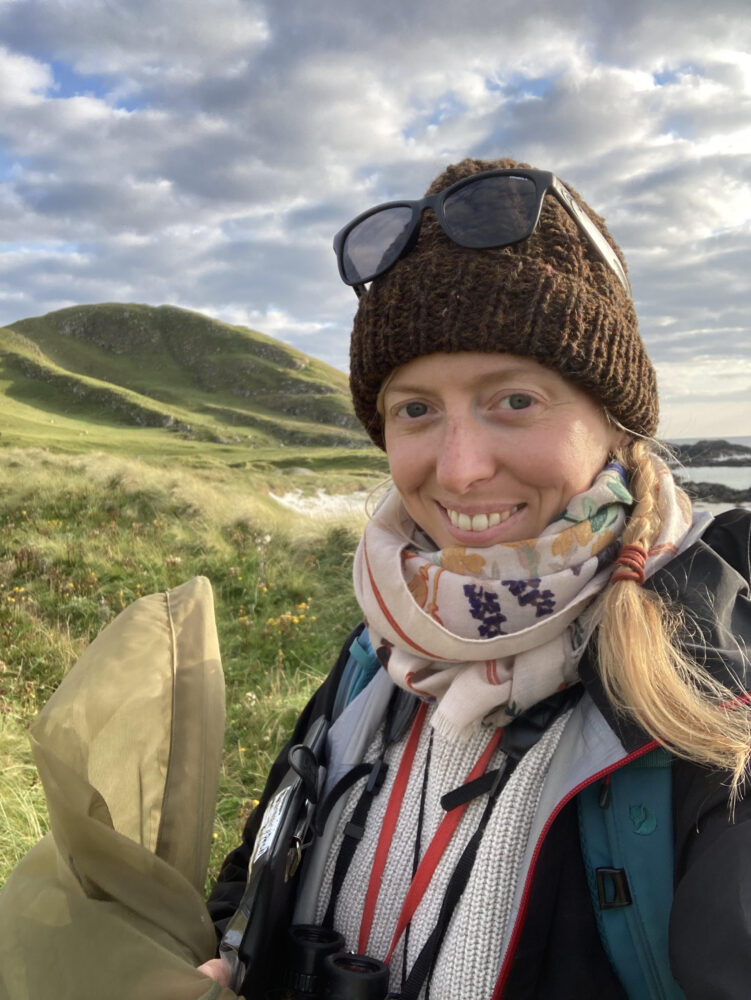 Person wearing a brown knit hat with sunglasses on top, bundled in a scarf and jacket, standing in a grassy landscape with rolling hills under a cloudy sky.