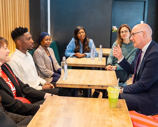Group of diverse professionals engaged in a discussion around wooden tables in a modern office setting.