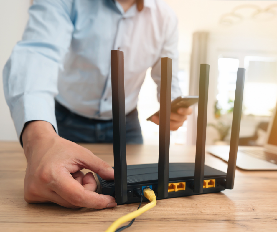 Person adjusting a black Wi-Fi router with four antennas on a wooden desk, connected by a yellow Ethernet cable in a bright office setting.