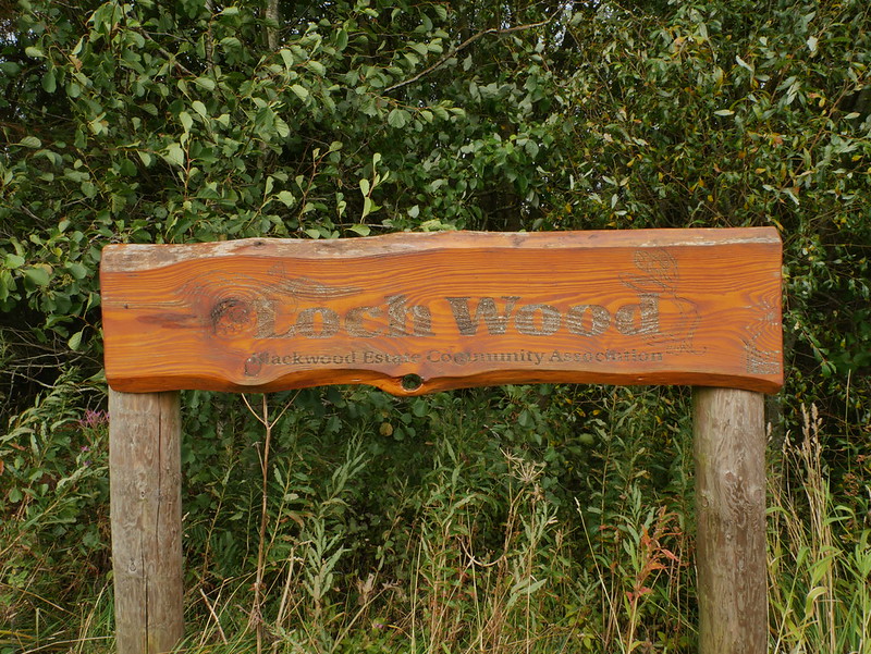 Wooden sign reading 'Park Word' with additional text about Backwood Estates Community Association, set against dense green foliage.