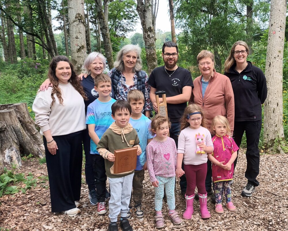 Group photo of four adults and seven children standing together in a wooded area with trees and leaf-covered ground.