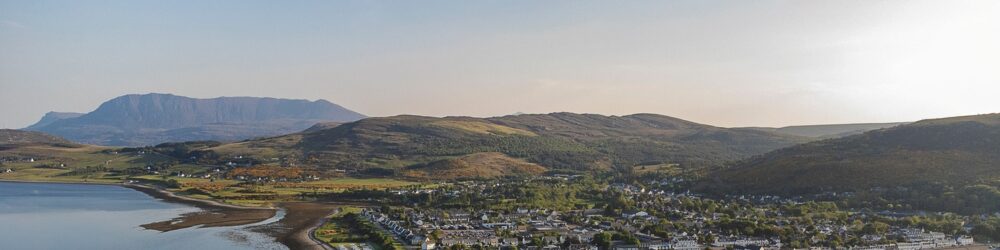 Wide-angle shot of a rural community with houses spread across grassy hills near a tranquil loch.