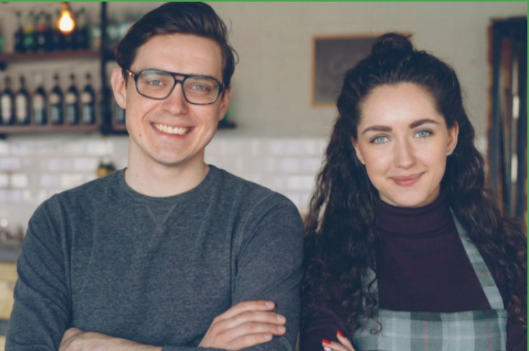 Smiling young man and woman standing with arms crossed inside a cozy café with wooden counters and shelves of bottles.