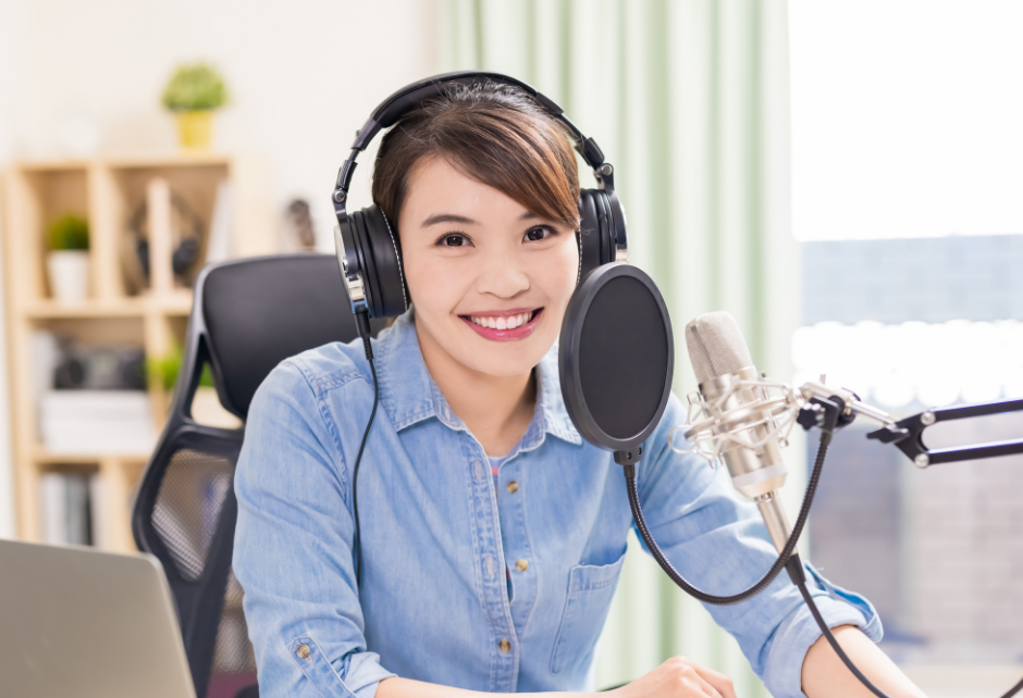 Woman in casual denim shirt hosting a podcast, using a pop filter microphone and laptop in a bright room.