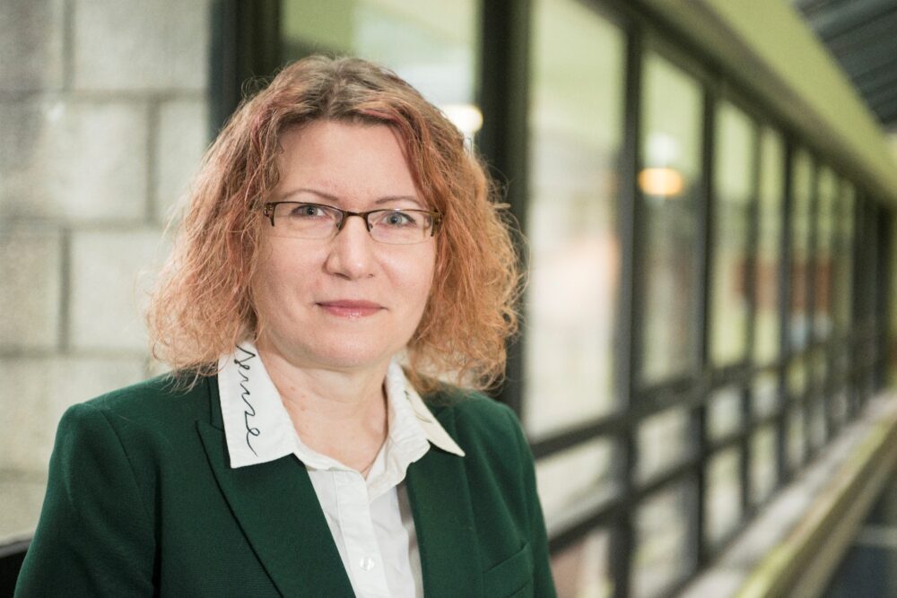 Professional woman with curly red hair and glasses wearing a green blazer and white shirt standing in a modern office corridor.
