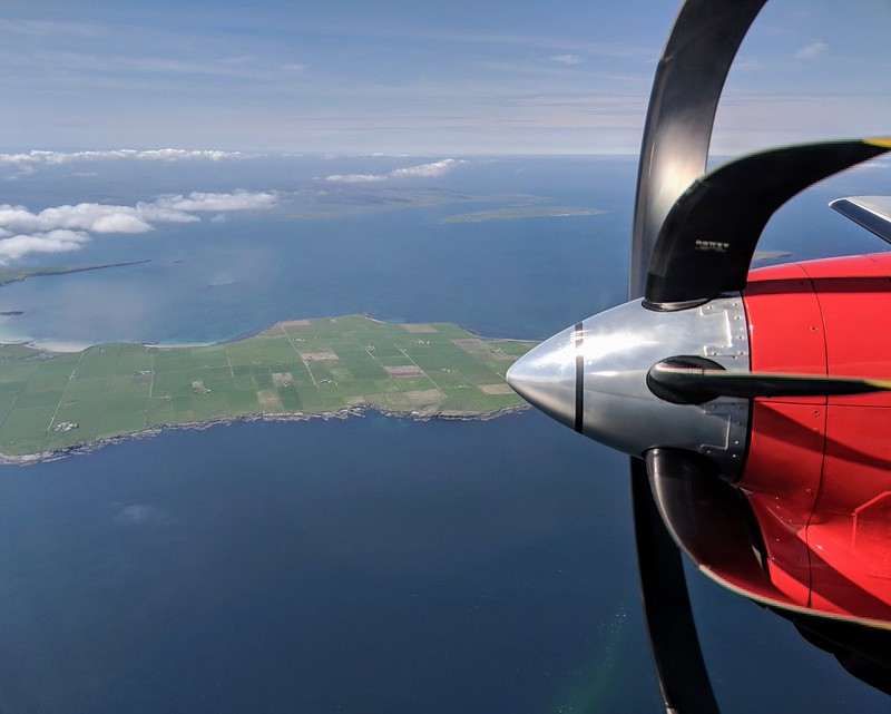 Aerial perspective of farmland on an island with a red airplane propeller in the foreground and calm sea below.