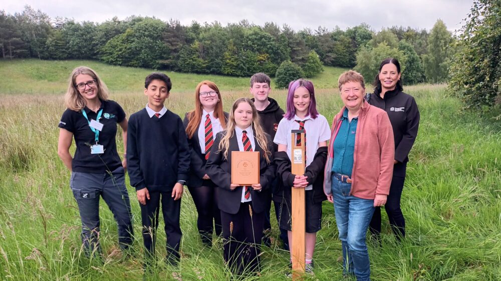 Group of seven students and two adults standing in a grassy field with trees in the background, some holding wooden plaques.
