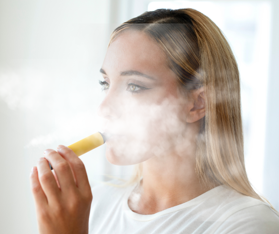 Young woman with blonde hair vaping indoors, exhaling a cloud of vapor from a yellow e-cigarette.